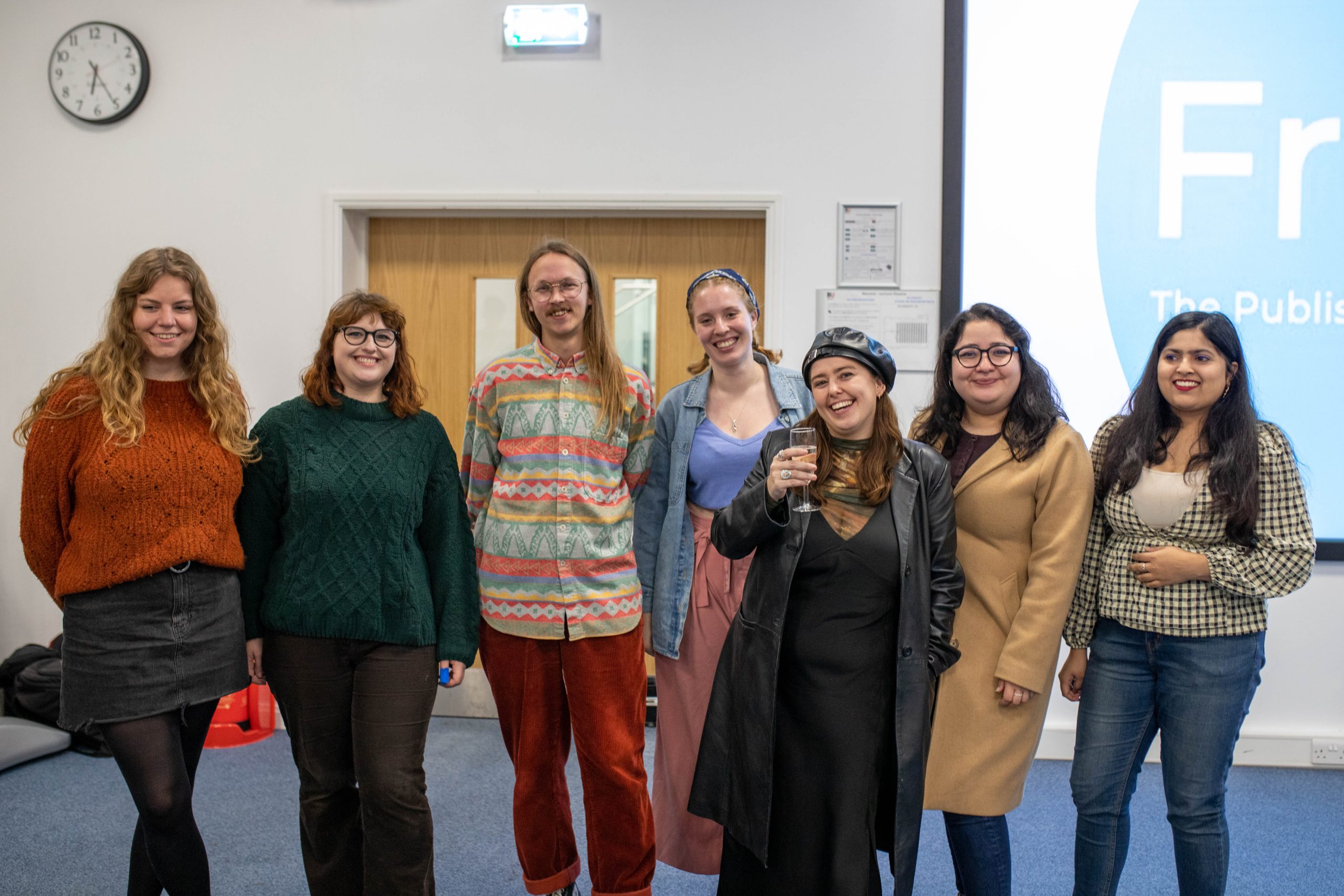 7 people stand in front of a wooden door and a projector showing the Freshers Publishing logo