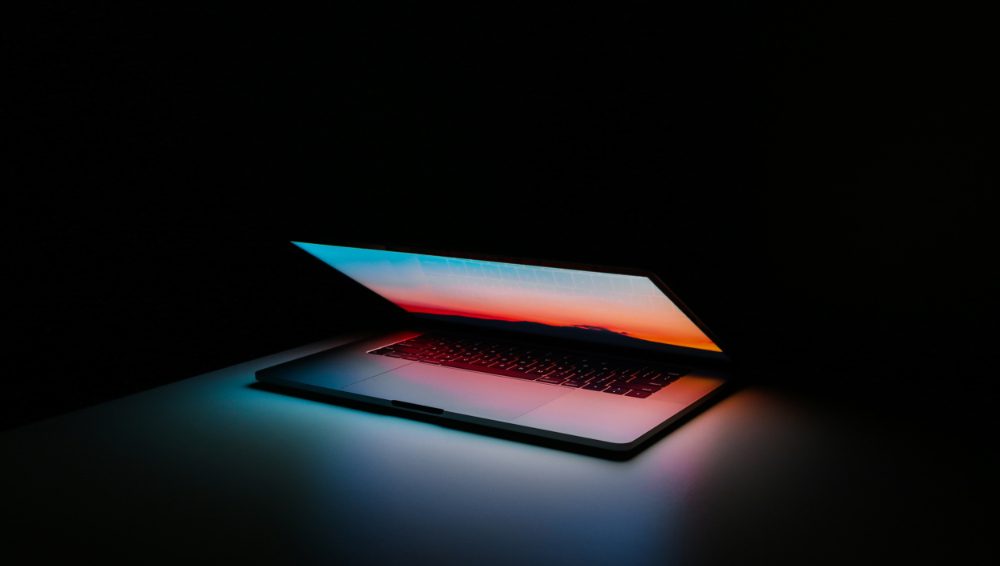 apple computer against a dark background. The computer is part way closed, the light of the home screen illuminates the keyboard in various colors.